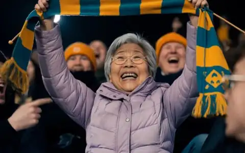 An elderly woman in a lilac coat lifts the teal and saffron scarf she has been knitting above her head and laughs as the fans around her cheer under the stadium floodlights